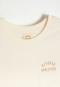 Kremowy bawełniany t-shirt z ryżowym dekoltem okrągłym i haftowanym napisem "Diverse Athletics" w zakrzywionym wzorze, umieszczonym na klatce piersiowej.