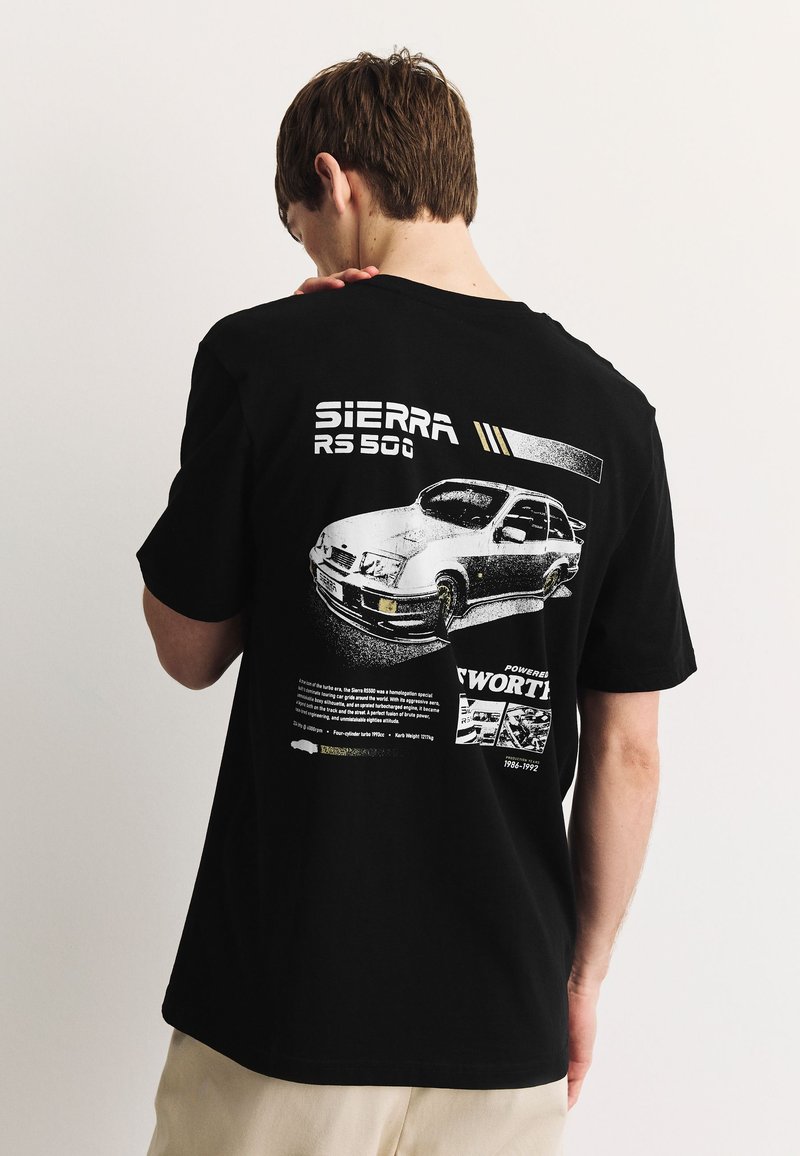 Noormees kannab musta T-särki, millel on valge pilt vintage Sierra RS 500 autost ja tekst seljal, seisab tavalise heleda tausta ees.
