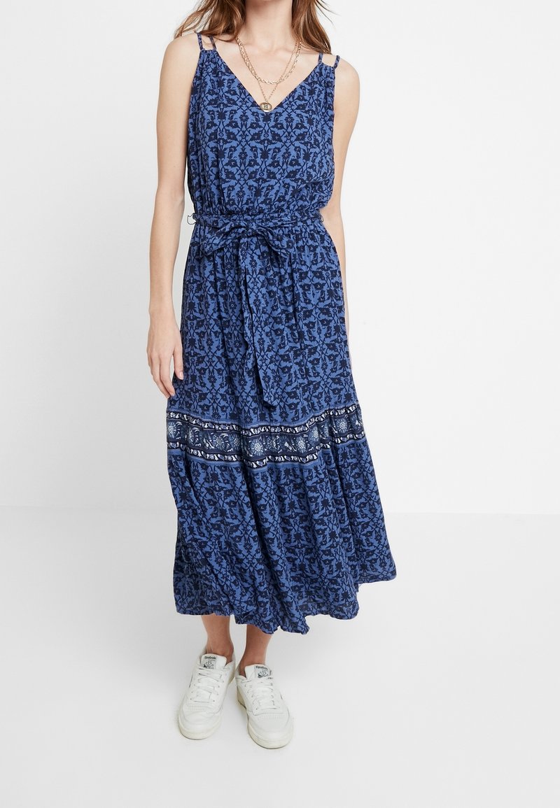 Robe longue à motif floral bleu avec une taille cintrée, un décolleté en V et une jupe fluide. Associée à des baskets blanches pour un look décontracté.