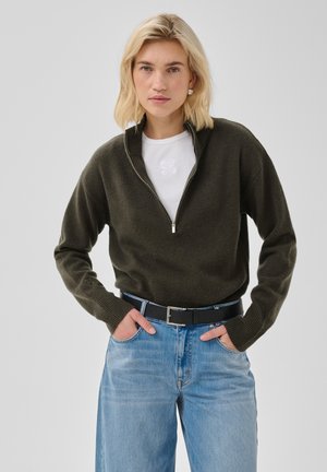 Femme blonde portant un pull vert foncé à demi-fermeture éclair, une chemise blanche, un jean bleu et une ceinture noire, debout les mains dans les poches devant un fond uni.