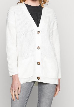 Woman wearing a white knitted cardigan with large buttons over a black top and gray jeans, standing against a plain background.