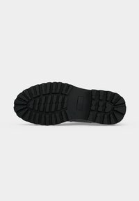Semelle de chaussure en caoutchouc noir avec des crampons profonds et larges pour une bonne traction, vue par dessous sur un fond clair uni.