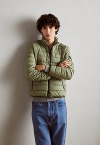 Giacca a vento color verde oliva chiaro, con zip frontale, polsini e orlo a coste, indossata sopra una maglietta grigia, abbinata a jeans in denim blu.