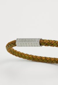 Zöld és arany szövetből készült fonott karkötő, ezüst színű fém címkével, amelyen a "GUESS" felirat látható. Az ékszer felülete textúrázott.