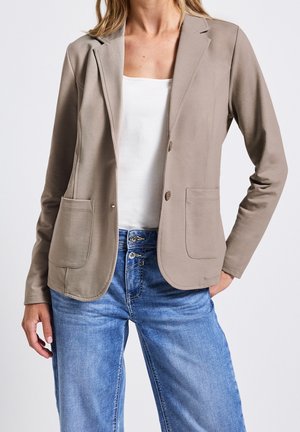 Vrouw met beige blazer met twee voorzakken over een witte top en blauwe spijkerbroek, poserend met één hand in de zak tegen een effen achtergrond.