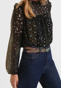Frau in einer schwarzen durchsichtigen Bluse mit floralem Muster, langen Ärmeln, hohem Kragen, glitzerndem Gürtel und dunkelblauen hoch-taillierten Jeans.