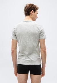 Homme aux cheveux bouclés portant un t-shirt gris clair à manches courtes et un short noir, debout de dos sur un fond uni.