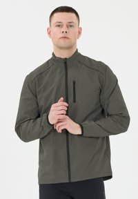 Ung man med kort hår, iförd en olivgrön jacka med dragkedja, som justerar ett handled mot en enkel vit bakgrund.