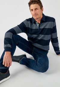 Gestreiftes Polohemd in Marineblau und Grau, mit langen Ärmeln und kragen mit Knopf. Tragen mit dunklen Jeans aus Denim und Marineblauen Turnschuhen mit weißen Sohlen.