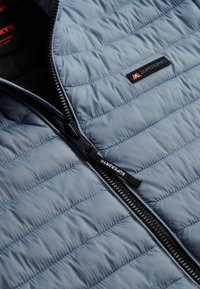 Jasnoniebieska pikowana kurtka z czarnym zamkiem częściowo otwartym, ukazująca czarną podszewkę z czerwoną logo Superdry.