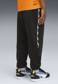 Kind trägt schwarze Hosen mit weißen Puma- und Hot Wheels-Logos, ein oranges T-Shirt und Puma-Sneaker mit blauen und gelben Akzenten.