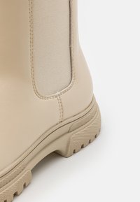 Botte cheville beige avec une tige synthétique lisse, un panneau latéral élastique et une semelle en caoutchouc texturée avec un épais relief.