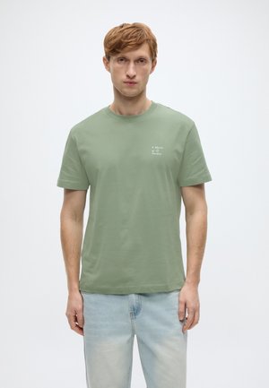 Olivgrünes Baumwoll-T-Shirt mit kurzen Ärmeln und Rundhalsausschnitt, versehen mit einem dezenten weißen Motiv auf der linken Brust, kombiniert mit hellblauen Jeanshosen.