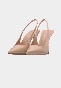 Beżowe czółenka na wysokim obcasie ze szpiczastym noskiem, wykonane z lakierowanej skóry, z cienkimi paskami i wąskim obcasem na białym tle.
