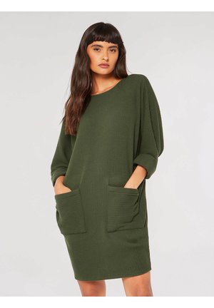 Groene ribgebreide jurk met een losse pasvorm, ronde halslijn en twee grote voorzakken. De lange mouwen zijn lichtjes opgerold.