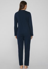 Žena stojící bosá, otočená zády, oblečená v tmavě modrém dlouhém rukávu a odpovídajících kalhotách rovného střihu na jednolitém pozadí.