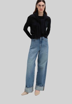 Czarna bluzka z długim rękawem, z dekoltem w łódkę i szalowym wzorem. Jasnoniebieskie jeansy z wysokim stanem, szerokimi nogawkami i podwiniętymi końcami. Beżowe buty z ostrym noskiem.