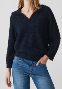 Pull en maille épaisse bleu marine avec un large col en V et un col. Manches longues et coupe décontractée. Associé à un jean en denim bleu clair.
