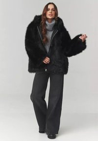 Czarna kurtka z imitacji futra z kapturem, z zapięciem na zamek, szerokimi rękawami, zestawiona z szarym golfem i czarnymi spodniami o szerokich nogawkach. Gładka tekstura.