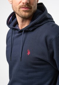 Marineblå hettegenser laget av tykk bomull, med et rødt brodert logo på brystet, justerbar hette og kontrasterende fargeaksenter på innsiden.