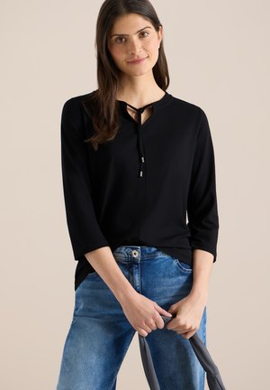 Femme portant un haut noir à manches longues avec un détail noué à l'avant, un jean bleu, tenant un tissu gris devant un fond beige.