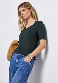 Tmavě zelený top s krátkým rukávem a výstřihem do V, zkombinovaný s modrými džíny. Modelka drží béžovou tašku a na ruce má zlatý náramek.