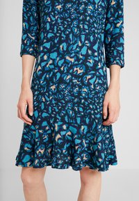 Klänning med blå och gröna blommönster, trekvartsärmar och utsvängd nederkant, tillverkad av mjukt tyg med en strukturerad yta.