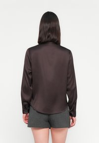 Blouse en satin marron à manches longues, col rond et poignets à boutons ; sa texture lisse et son léger éclat mettent en valeur son design élégant.