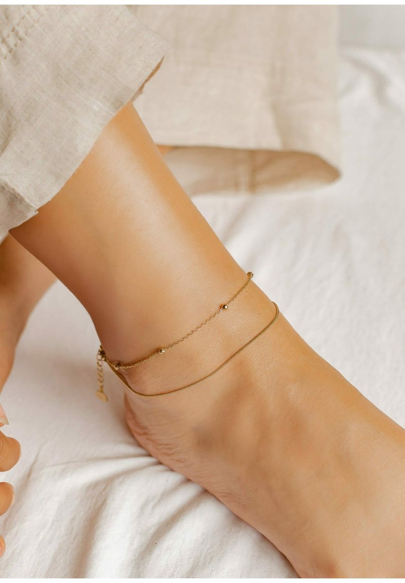 Tobillera en tono dorado con dos cadenas delicadas, una con pequeños acentos esféricos, descansando sobre un tobillo bronceado contra un fondo blanco texturizado.
