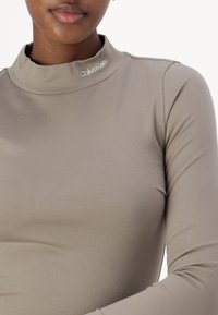 Długosleeve top w miękkim beżowym materiale z wysokim kołnierzem. Posiada logo Calvin Klein na dekolcie oraz dopasowany krój, co zapewnia elegancki wygląd.