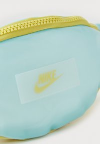 Rucsac fanny de culoare albastru deschis, realizat dintr-un material neted; are un fermoar galben și o etichetă albă cu un logo galben Nike.