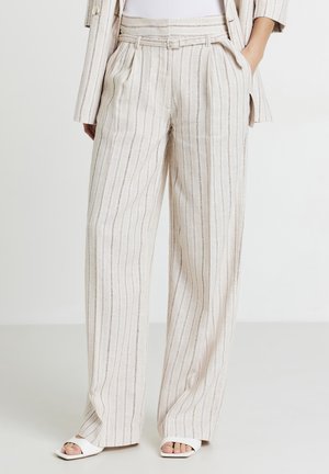 Donna che indossa pantaloni a gamba larga beige e bianchi a righe verticali con blazer abbinato e sandali bianchi con punta aperta, in piedi con le mani nelle tasche.