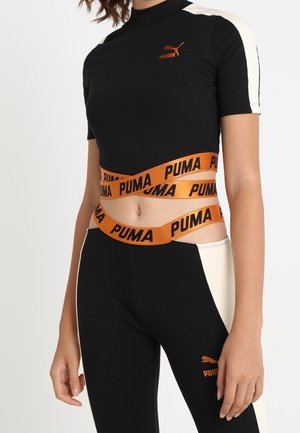 Musta ja kerma urheiluasu, jossa on lyhyt yläosa korkealla kauluksella ja oranssi brändin vyötärönauha. Leggingsien sivulla on kontrastivärinen paneeli.