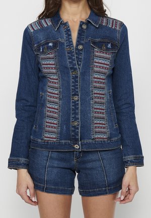 Vrouw draagt een donkerblauw spijkerjack met meerkleurige geweven patronen op de voorpanelen en bijpassende spijkershorts.