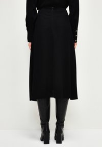 Jupe midi noire avec une texture lisse, une taille ajustée et une fermeture éclair dissimulée à l'arrière. Associée à des bottes noires montantes.
