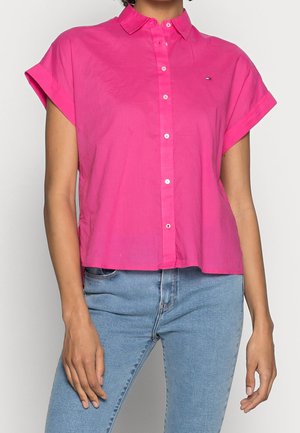 Nainen, joka pukeutuu kirkkaan vaaleanpunaiseen lyhythihaiseen napilliseen paitaan, jossa on pieni logo rinnassa, ja vaaleansinisiin farkkuihin.