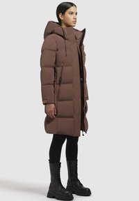 Pruun puffitakk, millel on kapuuts, pikkus, tikandite tekstuur, lukuga taskud ja mustad aktsendid. Kantud mustade retuuside ja saapade peal.