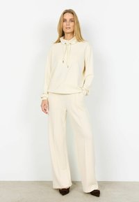 Krémovo sfarbená mikina s vysokým golierom a šnúrkou, v kombinácii so širokými nohavicami; obe majú jemnú textúru a voľný strih.