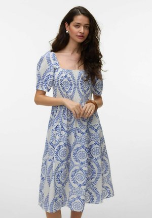 Kvinde med langt mørkt hår iført hvid midi-kjole med blå cirkulære blomsterprint, pufærmer og firkantet udskæring, stående mod hvid baggrund.