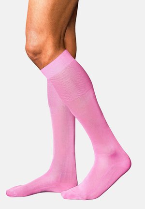 Beine mit kniehohen, hellrosa Kompressionsstrümpfen mit gerippten Bündchen vor schlichtem weißem Hintergrund.