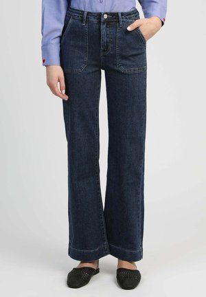 Personne portant un jean taille haute bleu foncé à jambes larges, une chemise à manches longues bleu clair et des chaussures noires tissées sans lacets.