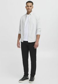 Männliches Outfit mit einem hell gestreiften Hemd mit Knöpfen, einem weißen T-Shirt, schwarzen Hosen und schwarzen Sneakern mit weißen Sohlen.