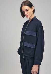 Giubbotto bomber navy con zip frontale, due grandi tasche a patta, polsini a costine e orlo elastico. Tessuto liscio con finitura opaca.