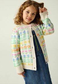 Strickjacke in Creme mit mehrfarbigem Blumenmuster; mit gerippten Bündchen und Knöpfen in Pink, Orange und Blau; kombiniert mit einem Jeanskleid.