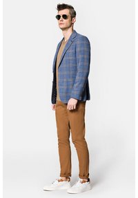 Niebieska marynarka w kratę z brązowymi akcentami, zestawiona z beżowym T-shirtem, brązowymi spodniami slim-fit i białymi sneakersami. Gładkie okulary przeciwsłoneczne dopełniają wygląd.