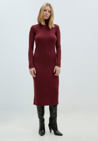 Merlot coltrui-jurk van zacht materiaal, aansluitende silhoët, reikt tot halverwege de kuit, gedragen met zwarte kniehoogte puntige laarzen.