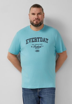 Homme avec barbe et cheveux attachés en arrière, debout les mains dans le dos, portant un T-shirt bleu clair avec le texte "EVERYDAY Rethink" et un jean bleu.