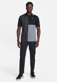 Svart och grå kortärmad polo skjorta med en färgblockdesign, tillsammans med svarta byxor och svartvita sneakers. Har en krage och logo.