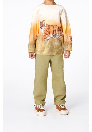 Kind trägt ein langärmeliges Hemd mit großem Tigerdruck, beige Hose und braune Sneakers mit weißen Sohlen und Schnürsenkeln.