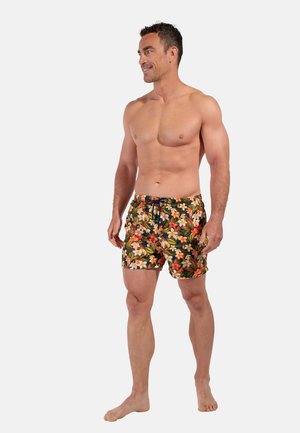 Oberkörperfreier Mann steht und trägt bunte, florale Badeshorts mit orange-, gelb- und grünfarbenen Mustern auf weißem Hintergrund.
